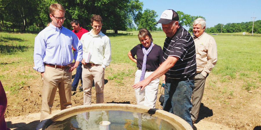 A man gestures toward a large cement watering trough while four people look on with interest.