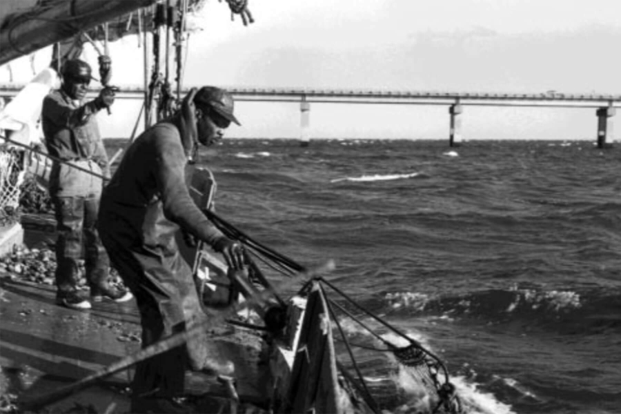 Two African-American watermen work on a boat in rough water. They are wearing rubber waders and working rigging and nets.